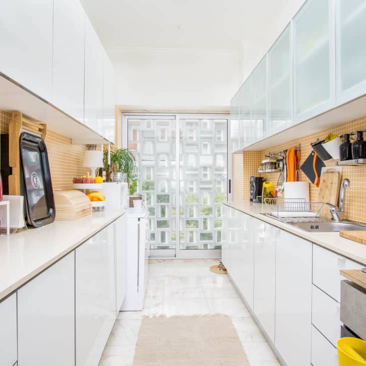 Modern galley kitchen with white cabinets, orange tile backsplash, colorful jars, and stainless steel appliances.