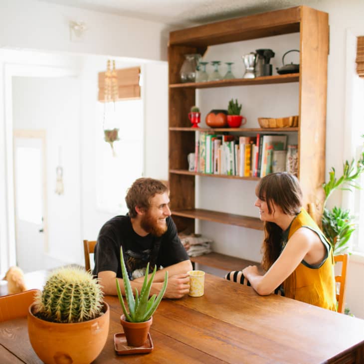 Couple sitting at a wooden table with potted cacti, surrounded by bookshelves and plants in a bright room.