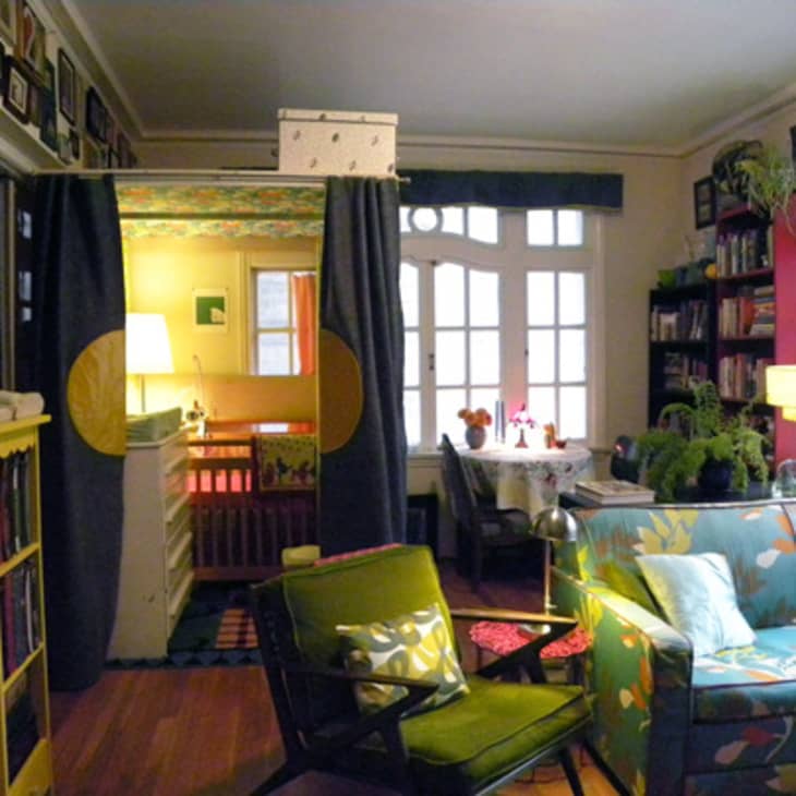 Living room with floral sofa, green armchair, bookshelves, and view into a nursery with crib and lamp.