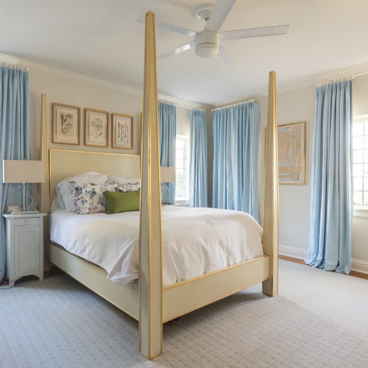 Four-poster bed with white linens, floral pillows, blue curtains, and botanical wall art in a bright bedroom.