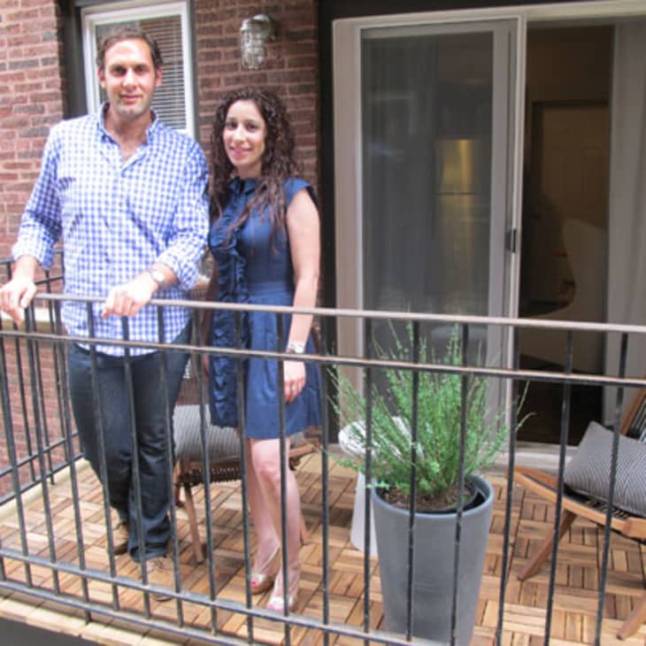 Two people standing on a small balcony with wooden flooring, potted plant, and sliding glass door.