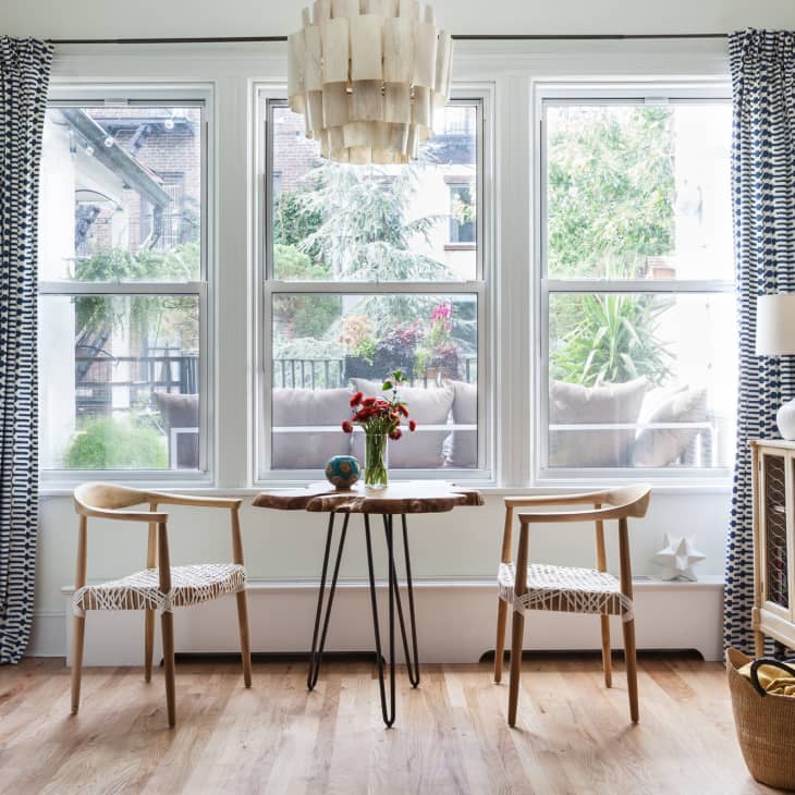 Two wooden chairs and a small table with flowers in a bright room with large windows and patterned curtains.