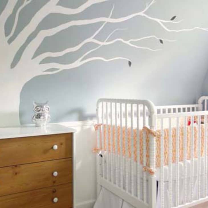 Nursery with white crib, wooden dresser, and tree mural on blue wall.