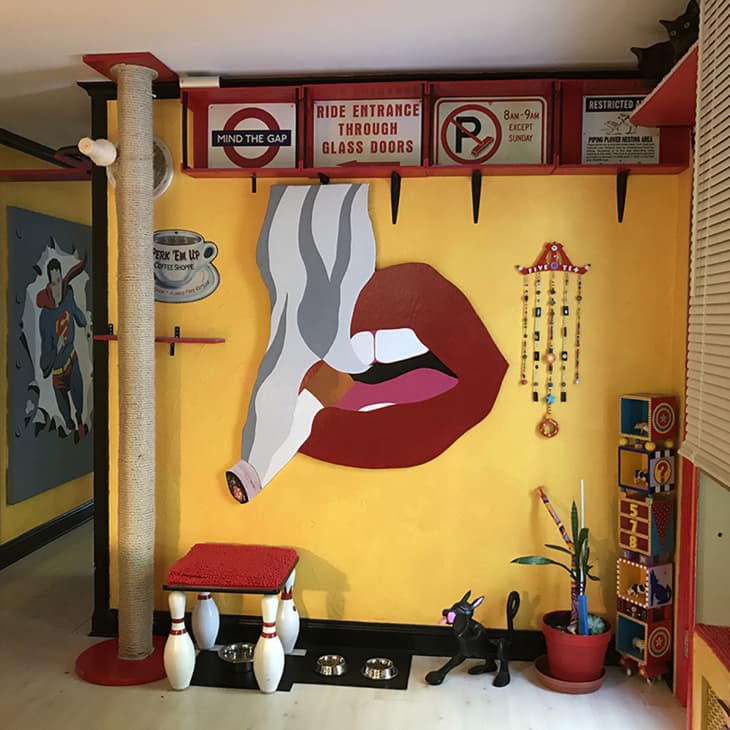 Colorful room with a large lips mural, retro signs, a cat scratch post, and eclectic decor items on a yellow wall.
