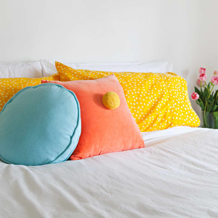 Bed with yellow polka dot pillows, blue and orange cushions, and pink tulips in a vase on a white nightstand.