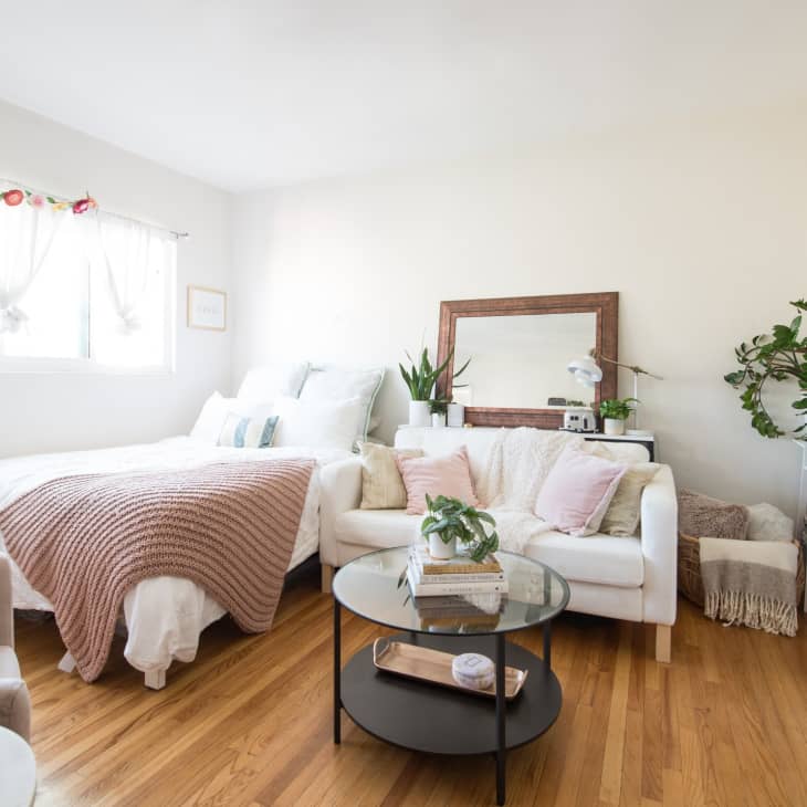Cozy bedroom with a white bed, pink throw, sofa, plants, and a mirror on a wooden floor.