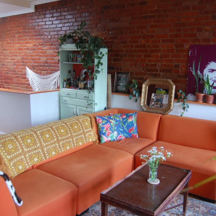 Orange sectional sofa with colorful pillows, brick wall, plants, and a vintage cabinet in a cozy living room.