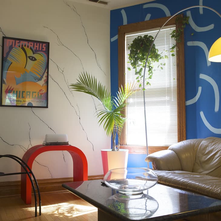 Living room with beige sofa, red table, Memphis poster, and blue accent wall with abstract patterns.