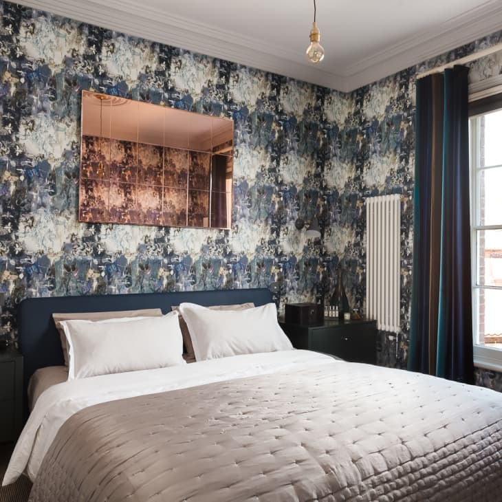 Bedroom with floral wallpaper, a double bed with white pillows, a quilted bedspread, and a large window with dark curtains.