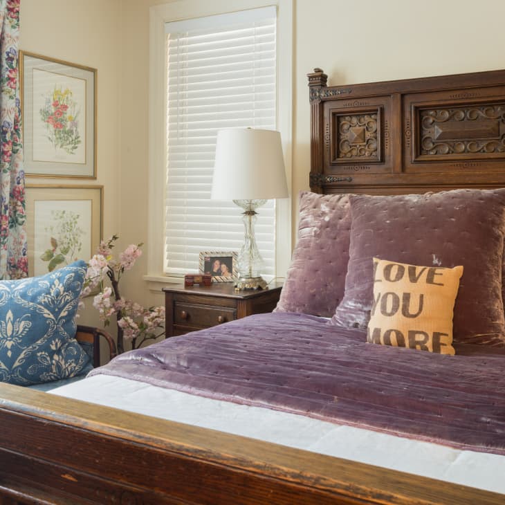 Ornate wooden bed with purple velvet bedding, "Love You More" pillow, floral curtains, and framed botanical prints.