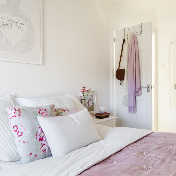 Cozy bedroom with floral pillows, pink throw, wall art, and a lavender scarf hanging on the door.