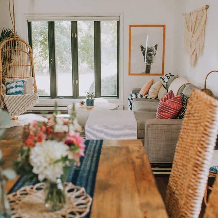 Cozy living room with a hanging chair, gray sofa, colorful cushions, and a wooden dining table with flowers.