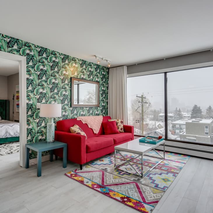 Living room with red sofa, tropical wallpaper, colorful rug, and large window view of snowy neighborhood. Bedroom visible through doorway.