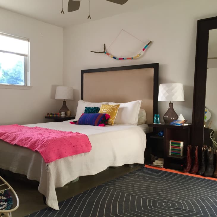 Bedroom with a beige headboard, colorful pillows, pink throw, large mirror, and cowboy boots lined up on the floor.