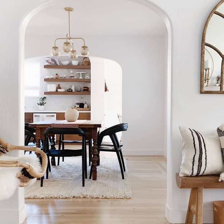 Arched entryway to dining room with wicker swing chair, wooden table, black chairs, and decorative pillows on a bench.