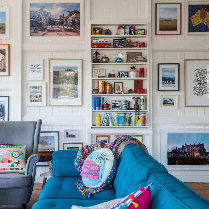 Living room with blue sofa, colorful pillows, gray armchair, and wall art gallery featuring framed photos and bookshelves.