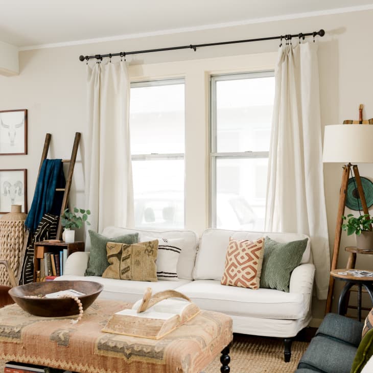 Cozy living room with white sofa, patterned cushions, wooden chairs, and a ladder shelf beside framed art and a floor lamp.