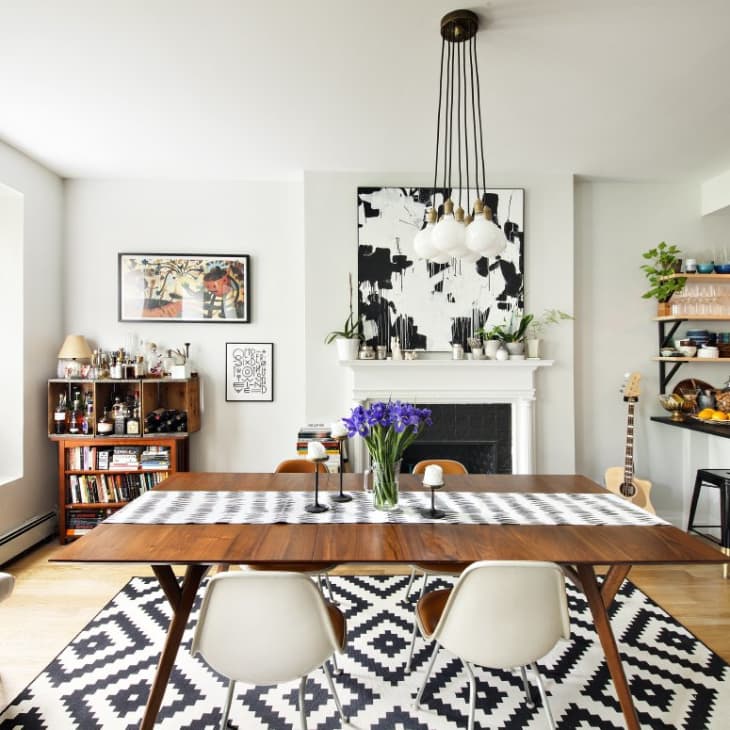 Dining room with wooden table, black and white rug, modern chandelier, and open shelves with plants and kitchenware.