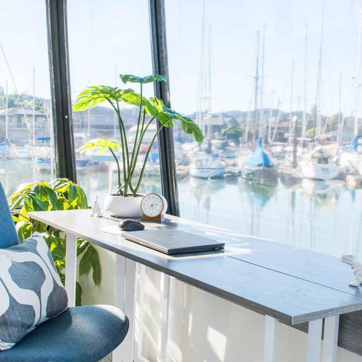 Home office with blue chair, desk, laptop, plant, and marina view through large windows.