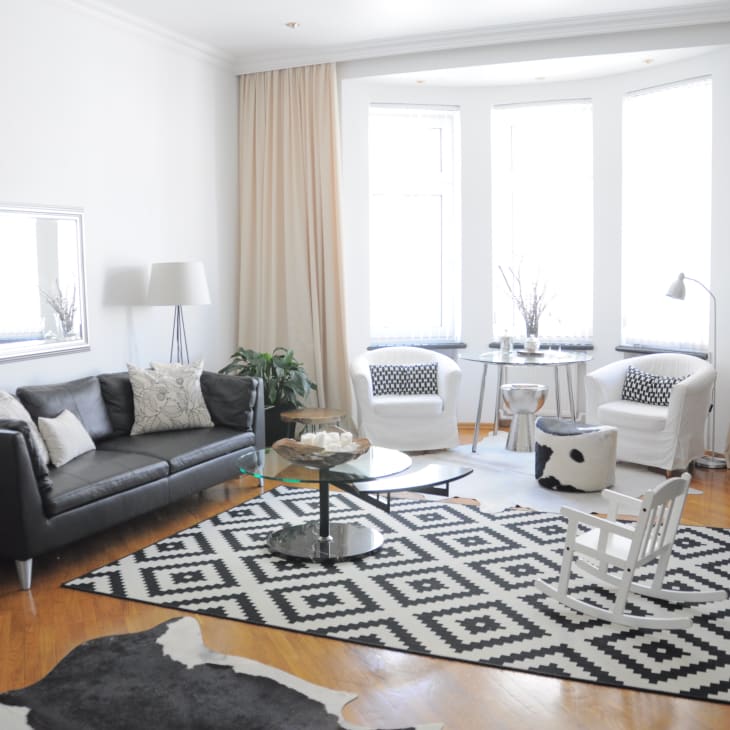 Modern living room with gray sofa, white armchairs, geometric rug, and large windows with beige curtains.