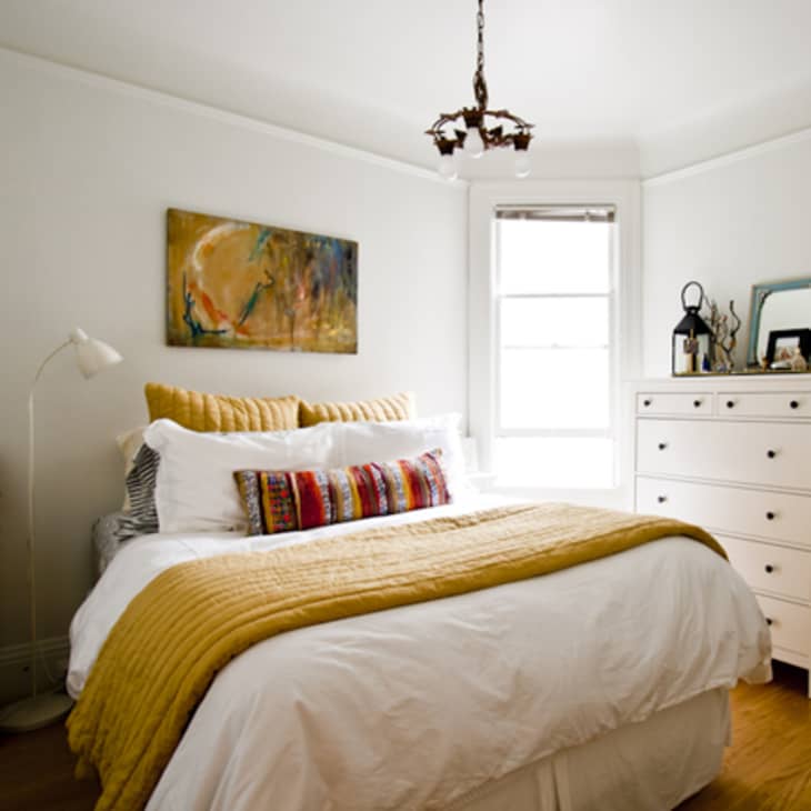 Cozy bedroom with a white bed, yellow accents, colorful pillows, a dresser, and abstract wall art.