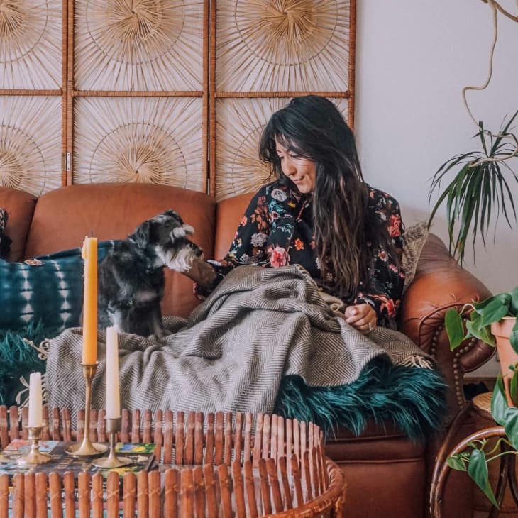 Woman sitting on a brown leather sofa with a dog, surrounded by plants and decorative pillows.