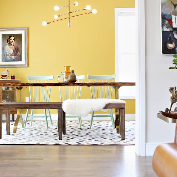 Dining room with yellow walls, wooden table, mint chairs, modern chandelier, and eclectic artwork.