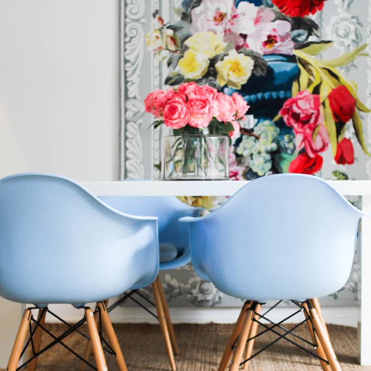 White dining table with blue chairs, pink flowers in a vase, and a colorful floral painting in the background.