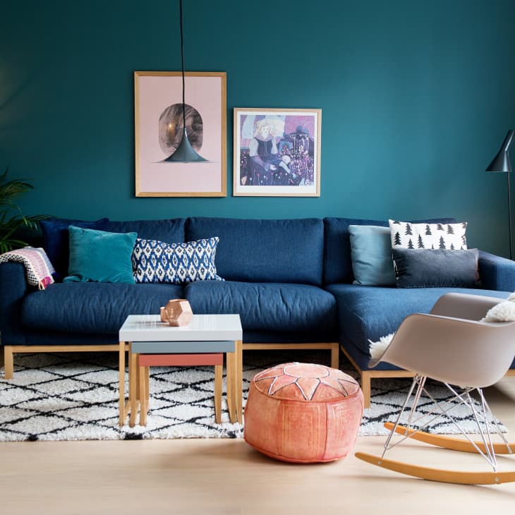 Modern living room with teal wall, blue sectional sofa, patterned cushions, pink pouf, and a beige rocking chair.