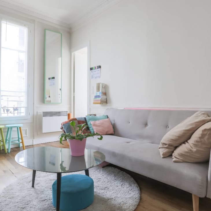 Bright living room with a gray sofa, colorful stools, a round glass coffee table, and a potted plant.