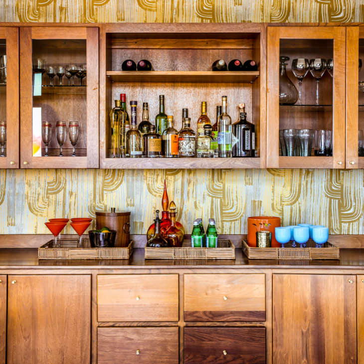 Wooden bar cabinet with glassware, liquor bottles, and decorative trays on a patterned wallpaper background.