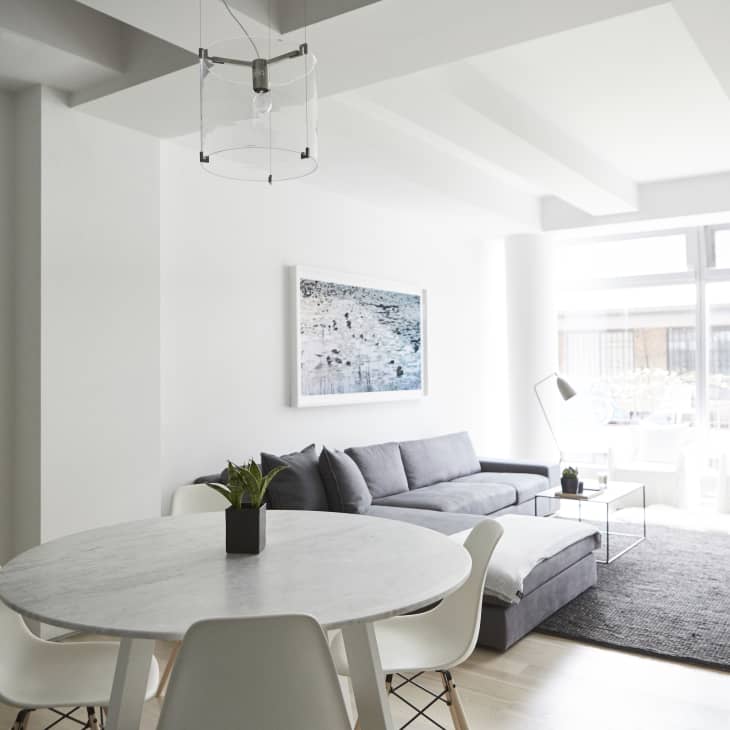 Modern living room with gray sectional sofa, round white dining table, white chairs, and large windows.