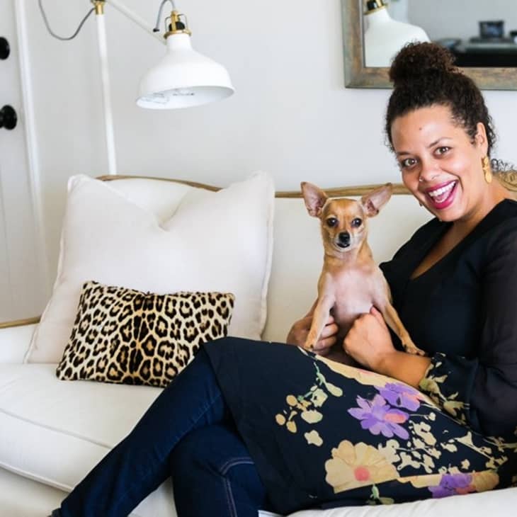 Woman sitting on a white sofa with a small dog, leopard print pillows, and a wall lamp.