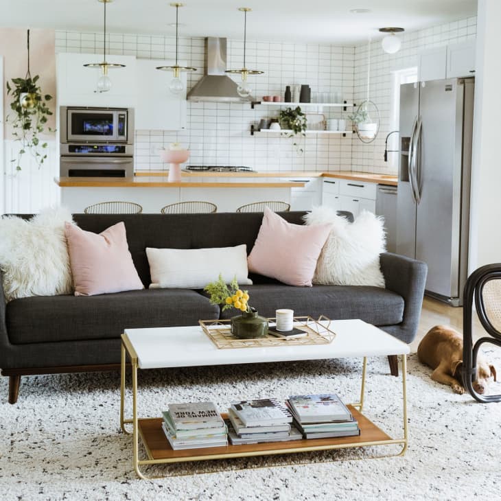Modern living room with dark sofa, pink and white pillows, white coffee table, and open kitchen in the background.