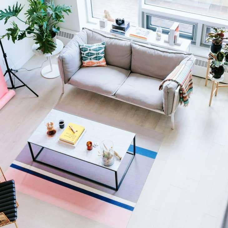 Living room with gray sofa, colorful pillows, coffee table with books, plants, and a pink backdrop.