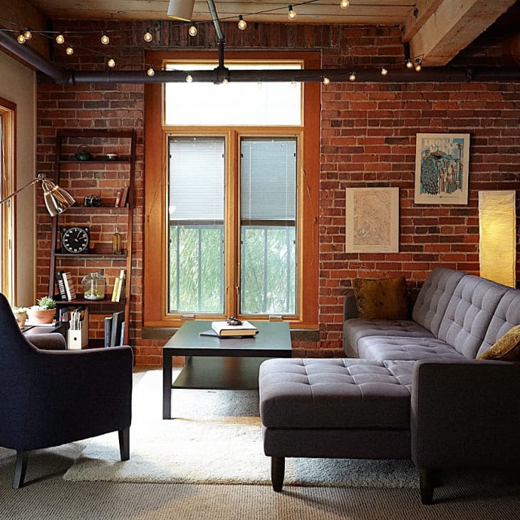 Loft living room with exposed brick, gray sectional sofa, armchair, bookshelves, and string lights.