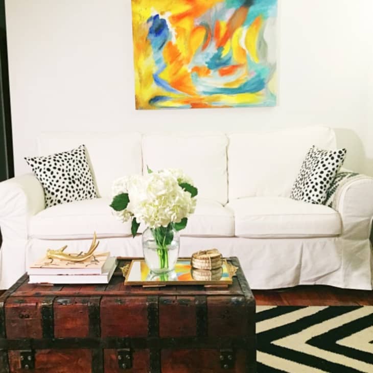 White sofa with polka dot pillows, abstract painting, wooden chest coffee table, and vase of white flowers.