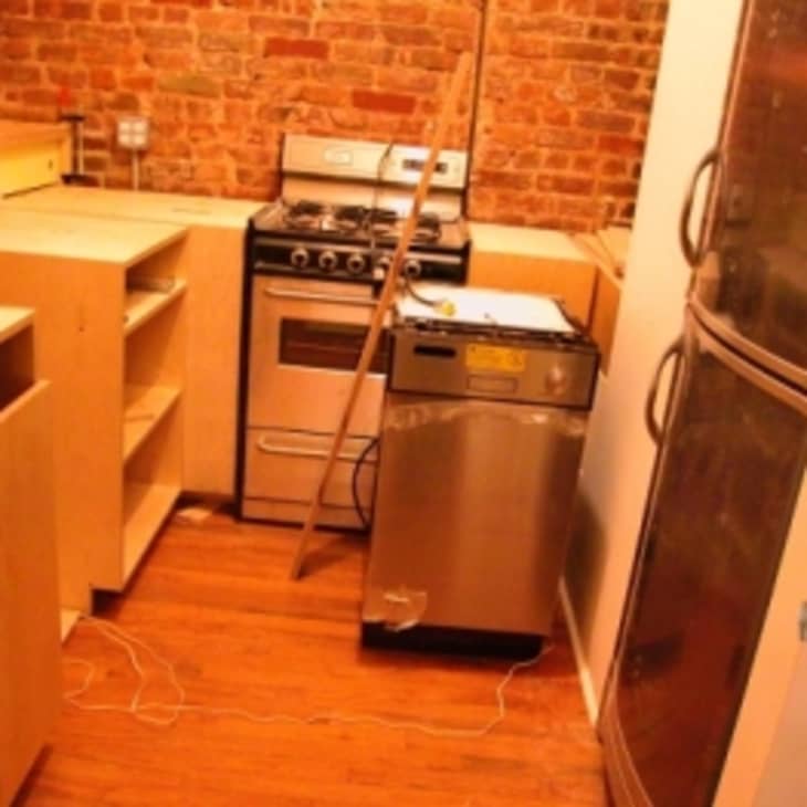 Small kitchen under renovation with exposed brick wall, stove, dishwasher, and unfinished wooden cabinets.