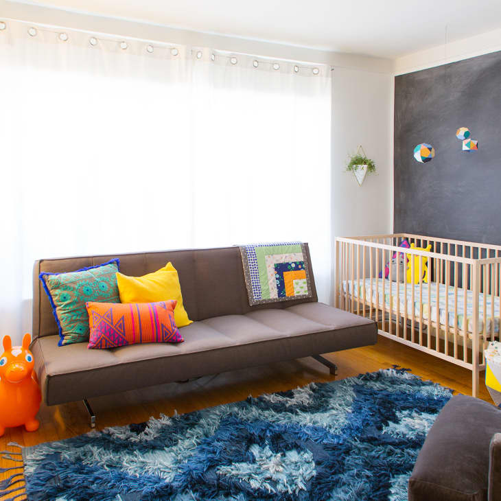 Colorful nursery with a gray sofa, vibrant pillows, wooden crib, blue rug, and playful decor on a chalkboard wall.