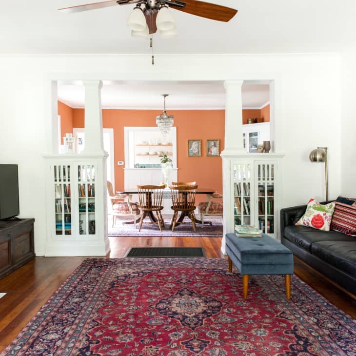 Living room with dark sofa, colorful cushions, red patterned rug, and view into dining room with orange walls and wooden table.