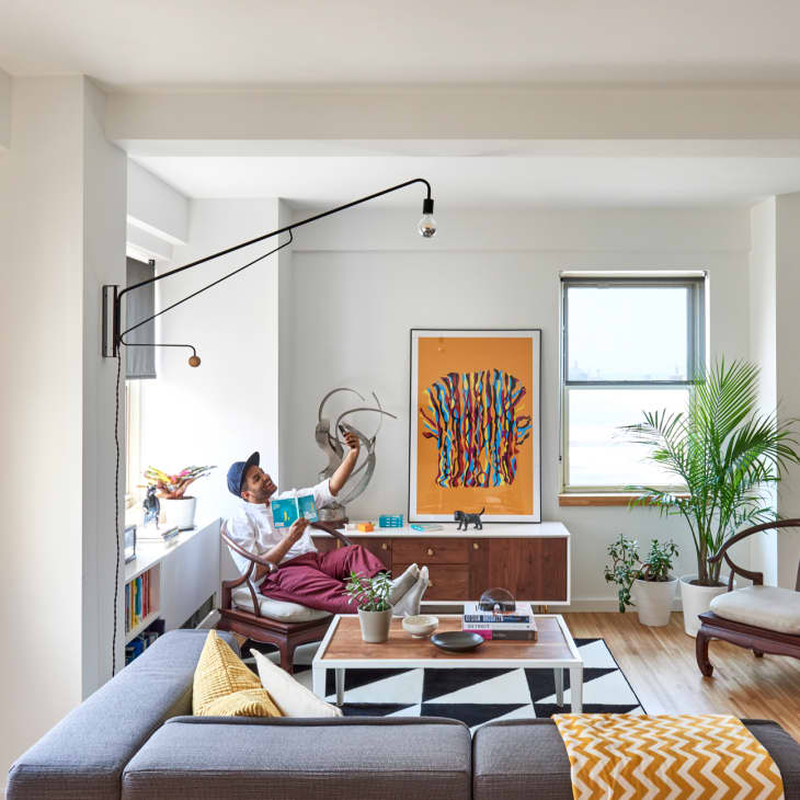 Modern living room with a gray sectional, geometric rug, wall art, and a person reading on a chair near a large plant.