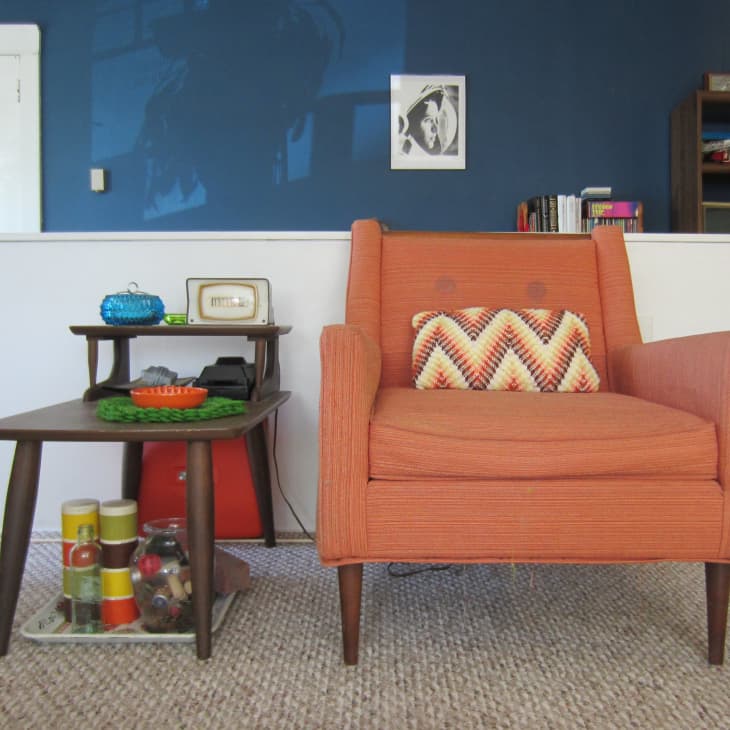 Orange armchair with chevron pillow beside a mid-century table holding a vintage radio and decor items, against a blue wall.