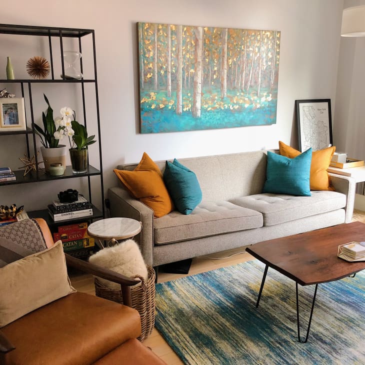 Cozy living room with a gray sofa, colorful pillows, abstract forest painting, and a wooden coffee table on a blue rug.