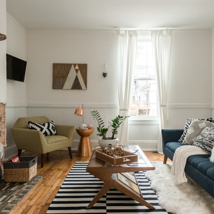 Cozy living room with blue sofa, patterned cushions, green armchair, striped rug, and decorative plants near a rustic fireplace.