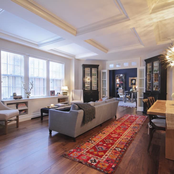 Open living and dining area with a gray sofa, red patterned rug, wooden table, and a starburst chandelier.