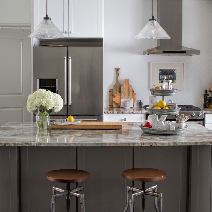 Modern kitchen with marble island, two stools, stainless steel fridge, pendant lights, and decorative cutting boards.
