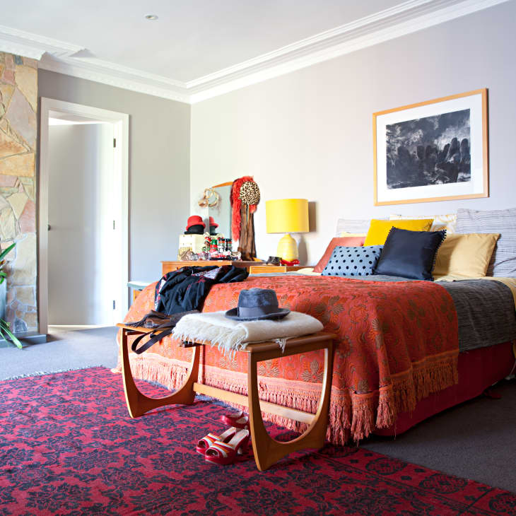 Bedroom with stone fireplace, red patterned rug, orange bedspread, yellow lamps, and wooden side tables.
