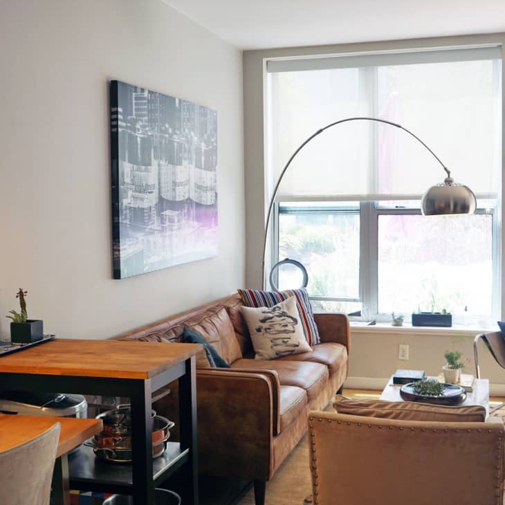 Cozy living room with brown leather sofa, modern art, arc floor lamp, and decorative pillows near a large window.