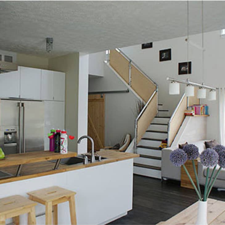 Modern kitchen and living area with white cabinets, wooden countertops, bar stools, staircase, and large windows.