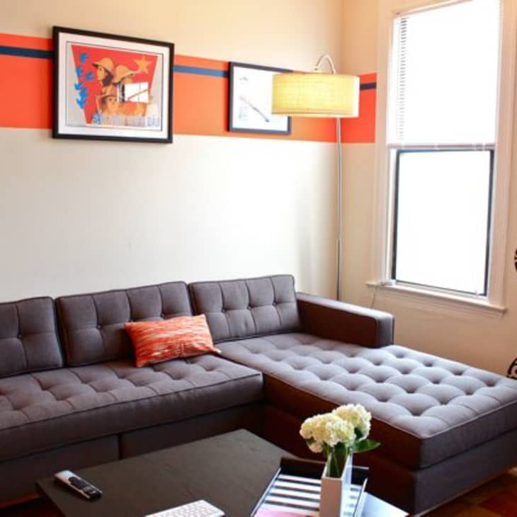 Gray sectional sofa with orange pillow, wall art, floor lamp, and patterned wallpaper in a cozy living room.
